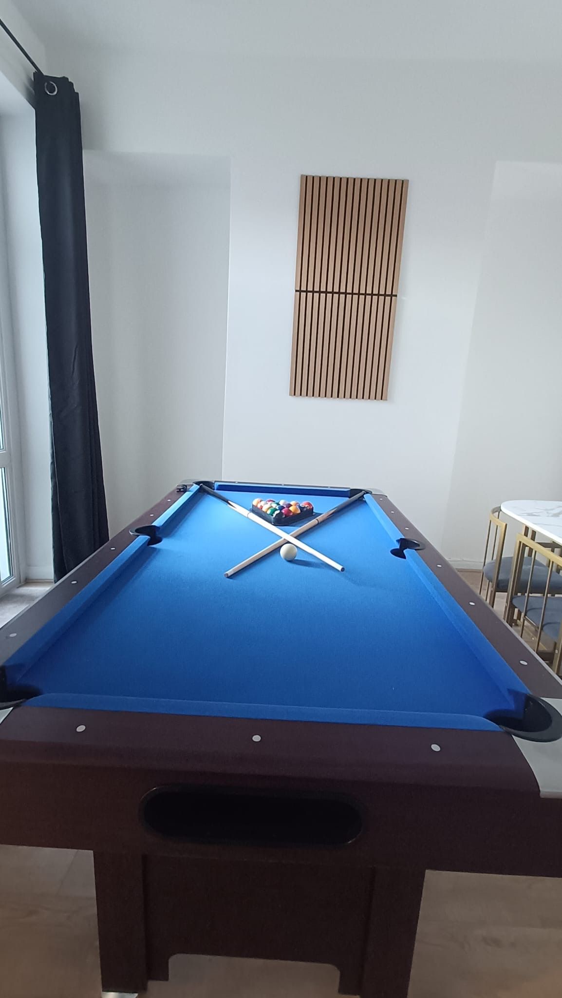 Una mesa de billar con bolas y tacos, una ventana con cortina y un mueble para colgar en la pared.