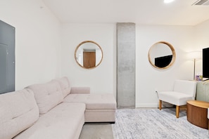 A modern living room with a sectional sofa, a small white chair, a round mirror, and a lamp on a side table. - Vibrant Japantown Townhome | Walk to Dining (San Jose)