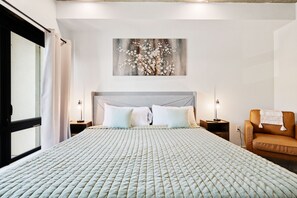 Ein Hotelzimmer mit einem großen Bett, einem Kopfteil, zwei Nachttischen, einem Sessel und einem an der Wand h ängenden Kunstwerk.