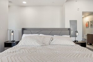 A bedroom with a large bed, two bedside tables with lamps, a grey headboard, and a door leading to another room. - Silicon Valley Basecamp | Townhome in Japantown SJ (San Jose)
