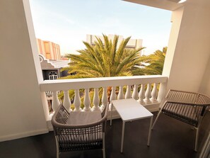 A balcony with a white railing, two wicker chairs, a small white table, and a view of palm trees and buildings. - Modern Downtown Loft Walk2Beach I CasaAmarilla304 (Oranjestad)