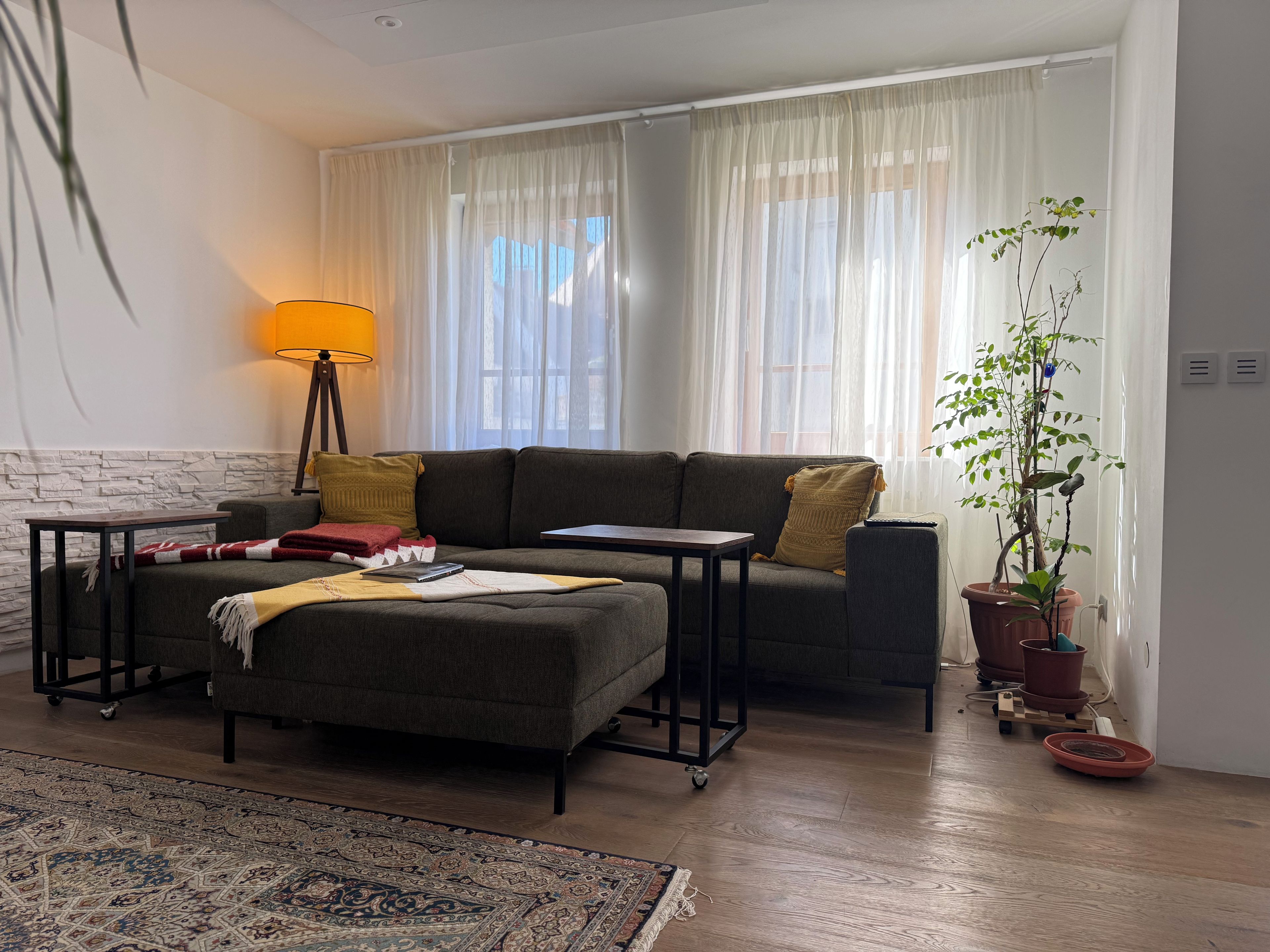 Una sala de estar con sofá, otoman, mesitas auxiliares, lámpara, planta en maceta y alfombra.