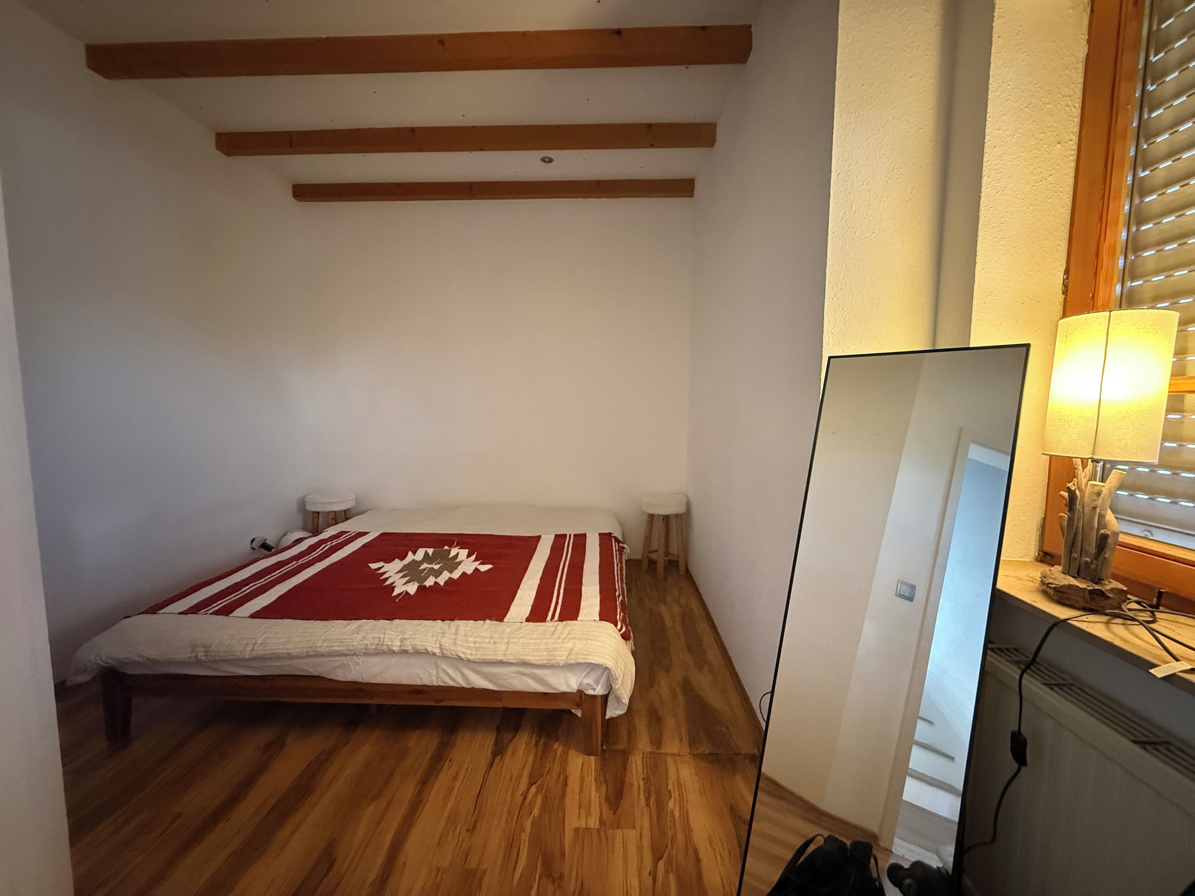 Ein Schlafzimmer mit einem Bett, Holzbalken an der Decke, einem Nachttisch, einer Lampe und einem Fenster mit Jalousien.