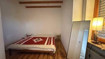 Ein Schlafzimmer mit einem Bett, Holzbalken an der Decke, einem Nachttisch, einer Lampe und einem Fenster mit Jalousien.