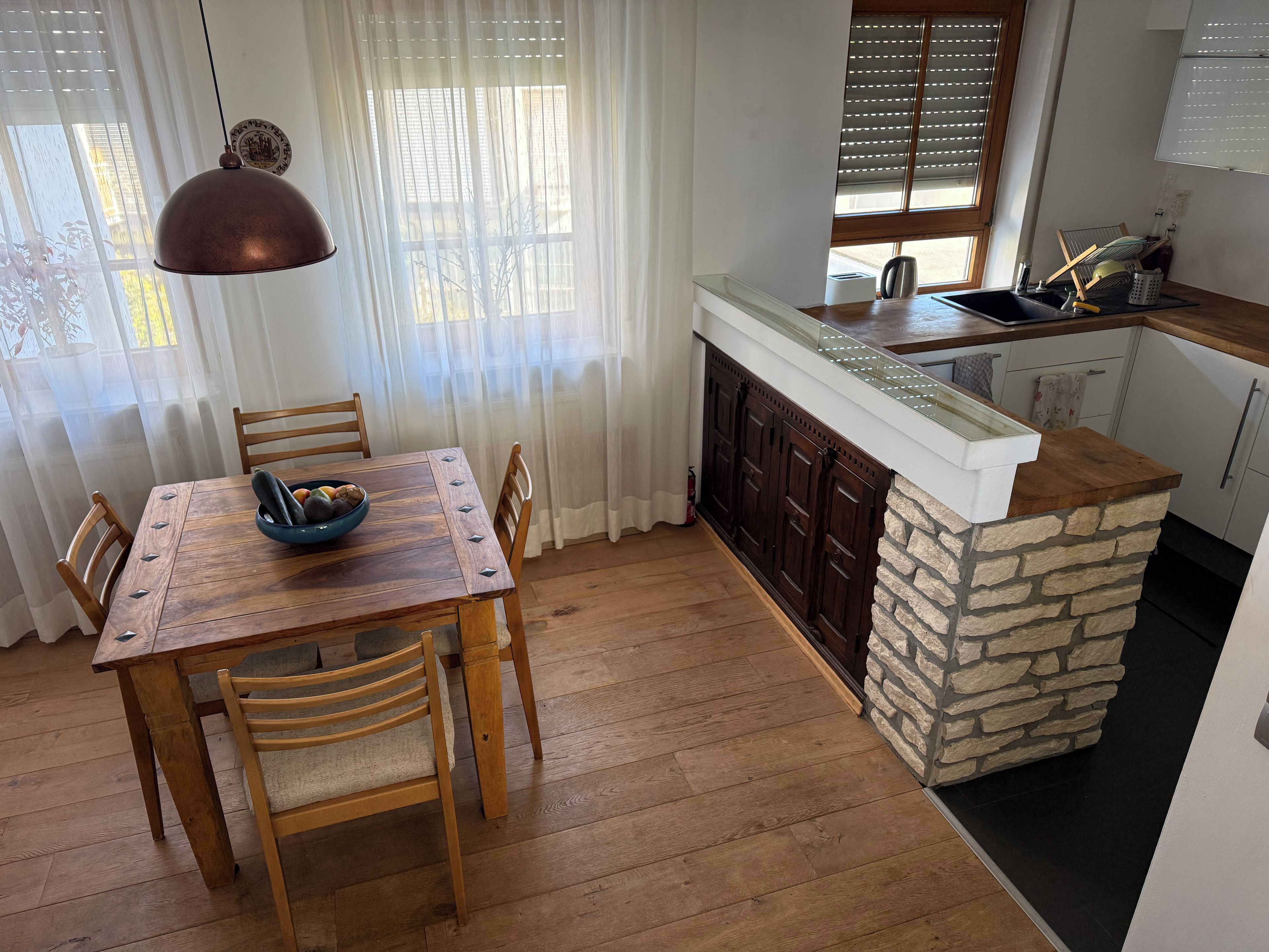 A dining area with a wooden table and chairs, a kitchen with a stone accent wall, and a window with blinds.