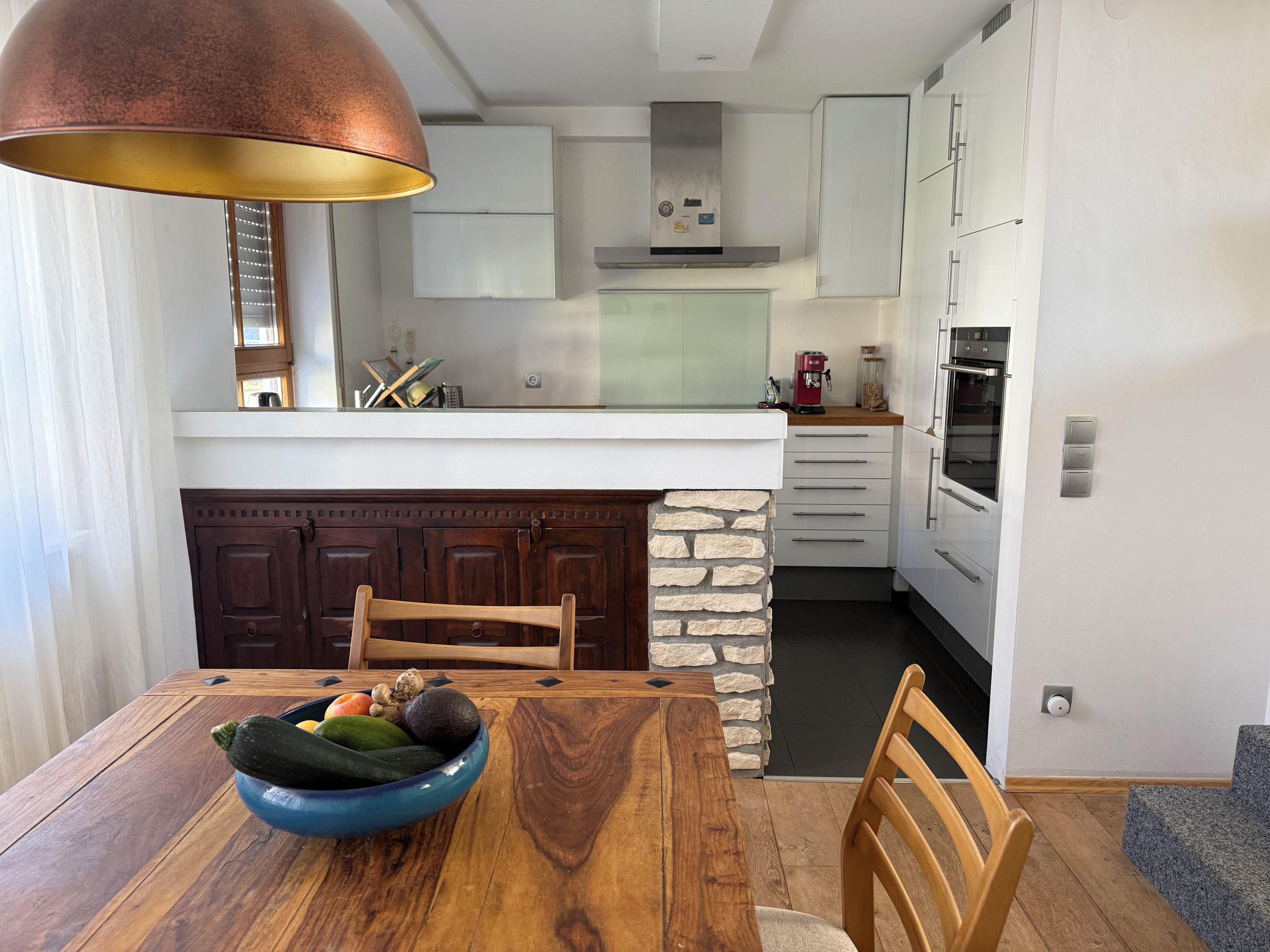 A modern kitchen with white cabinets, a stone accent wall, and a wooden dining table with a bowl of fruit.
