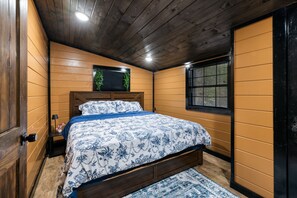 A bedroom with a wooden ceiling, a bed with a floral bedspread, a window with blinds, and a small plant on the wall. - Wood, Water & Wows:Loaded Cabin with GameRoom near Lake (Broken Bow)