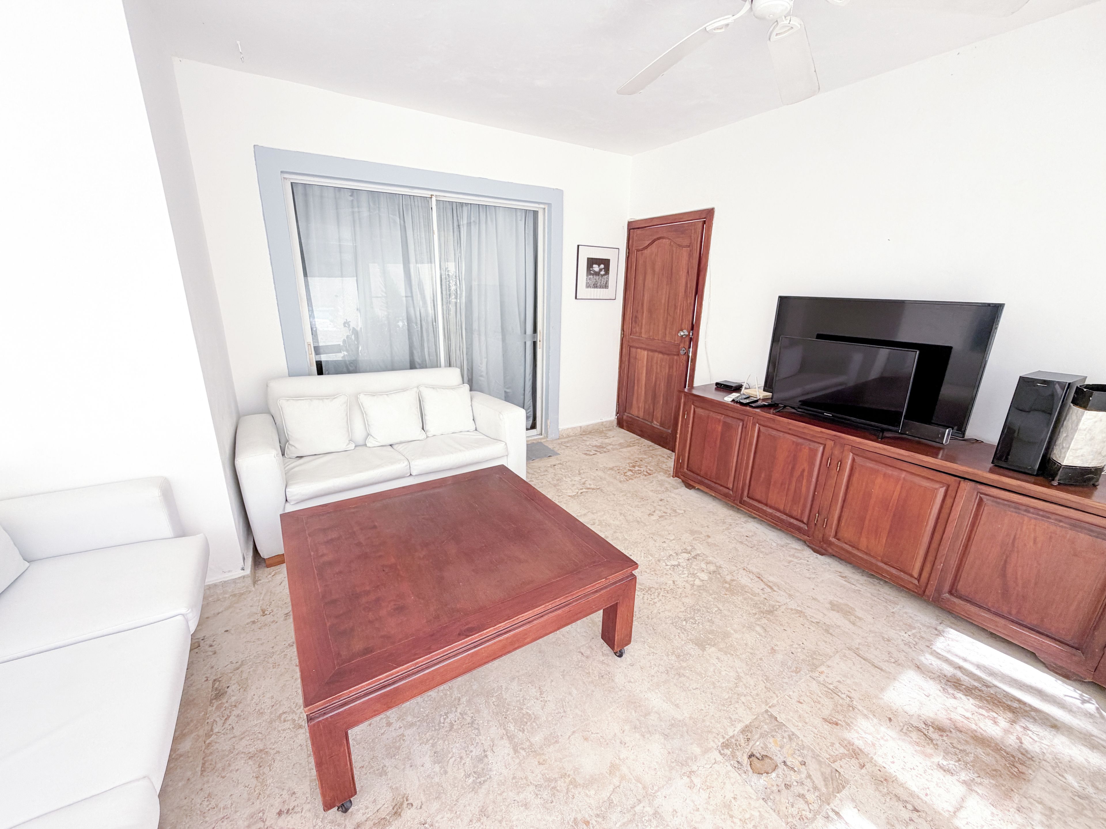 Un salón con un sofá blanco, una mesa de centro de madera, un televisor sobre un mueble de madera y una puerta corredera de cristal con cortinas.