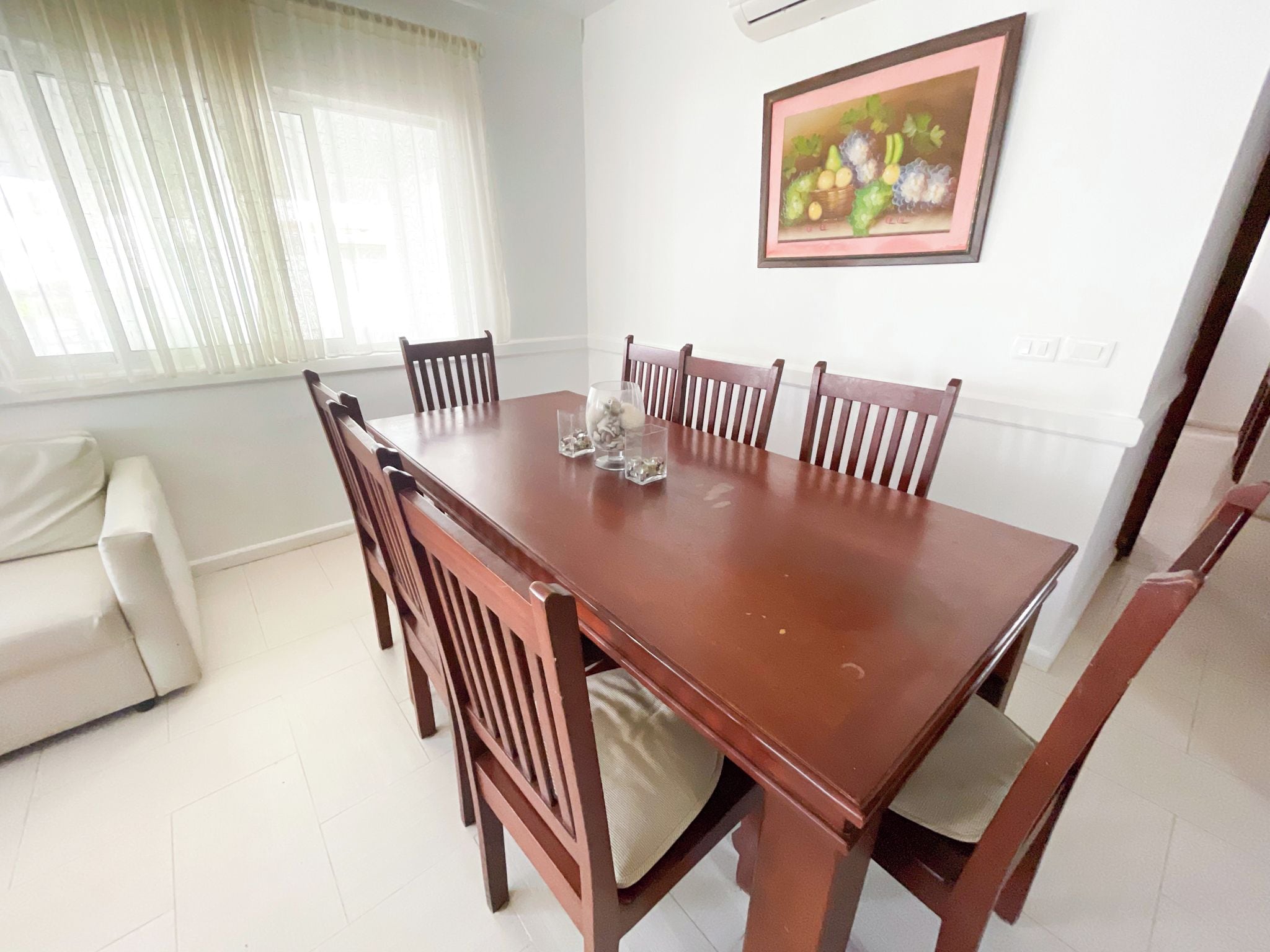 Un área de comedor con una mesa y sillas de madera, un sofá blanco y una ventana con cortinas translúcidas.