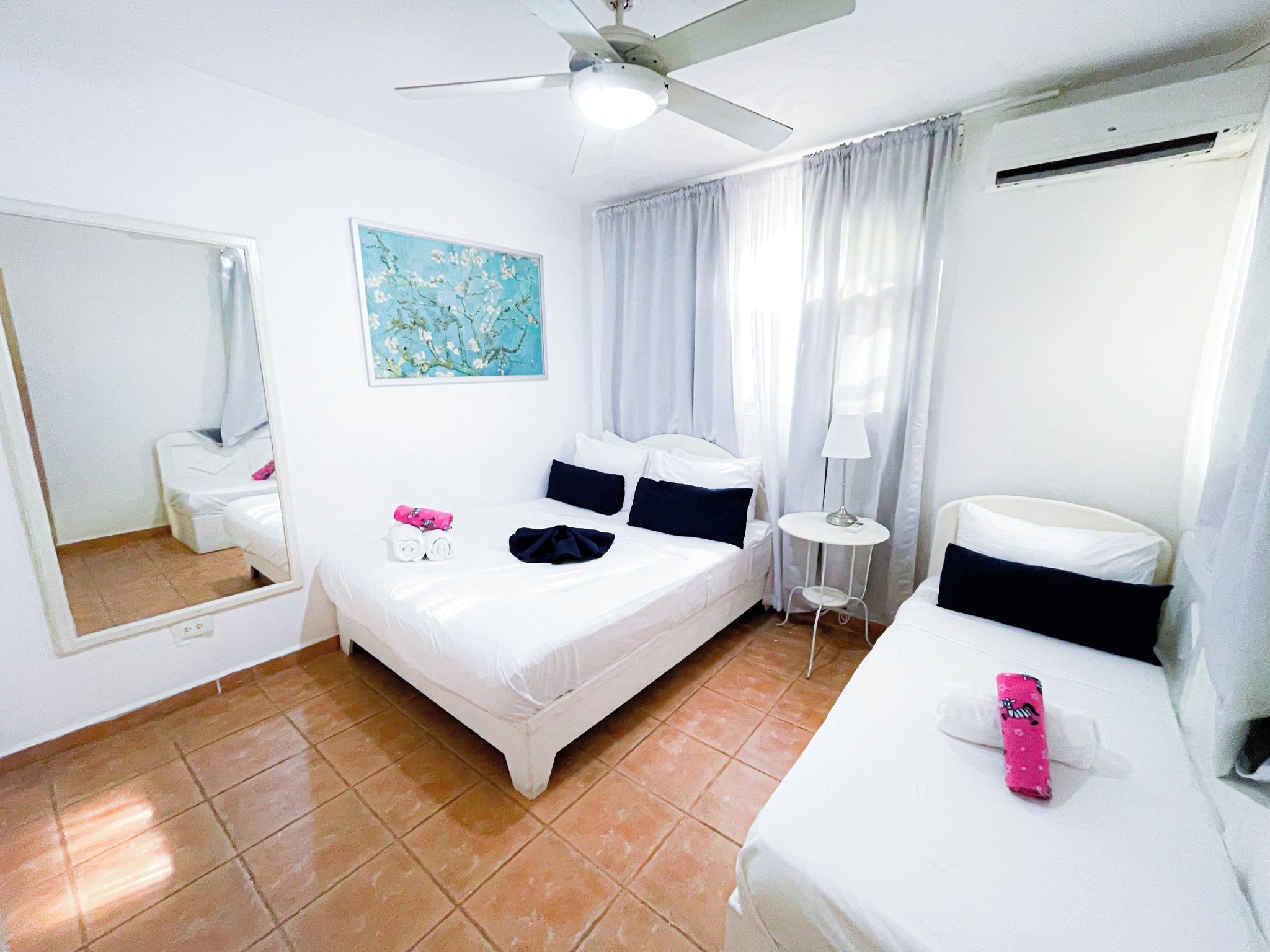 Une chambre d’hôtel avec un lit, une table de chevet, une lampe, un ventilateur de plafond, une fenêtre avec des rideaux et un tableau au mur.