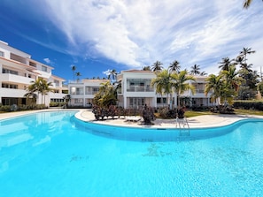A swimming pool surrounded by modern buildings with balconies, palm trees, and a clear sky. - VILLAS and APARTMENTS playa LOS CORALES (Punta Cana)