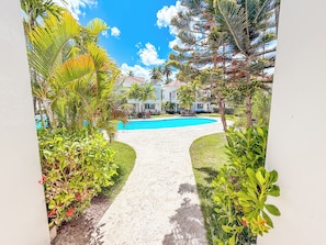 A pathway leading to a swimming pool surrounded by lush greenery and a building with a red-tiled roof. - VILLAS and APARTMENTS playa LOS CORALES (Punta Cana)