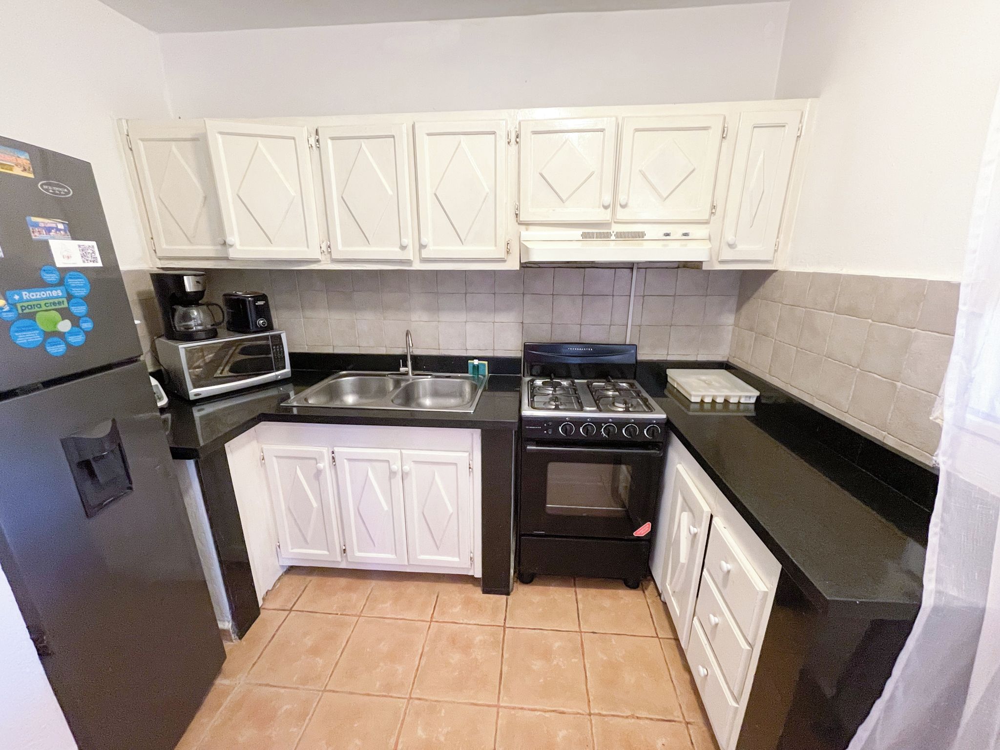 A compact kitchen with white cabinets, a black refrigerator, a microwave, a coffee maker, a gas stove, and a sink.
