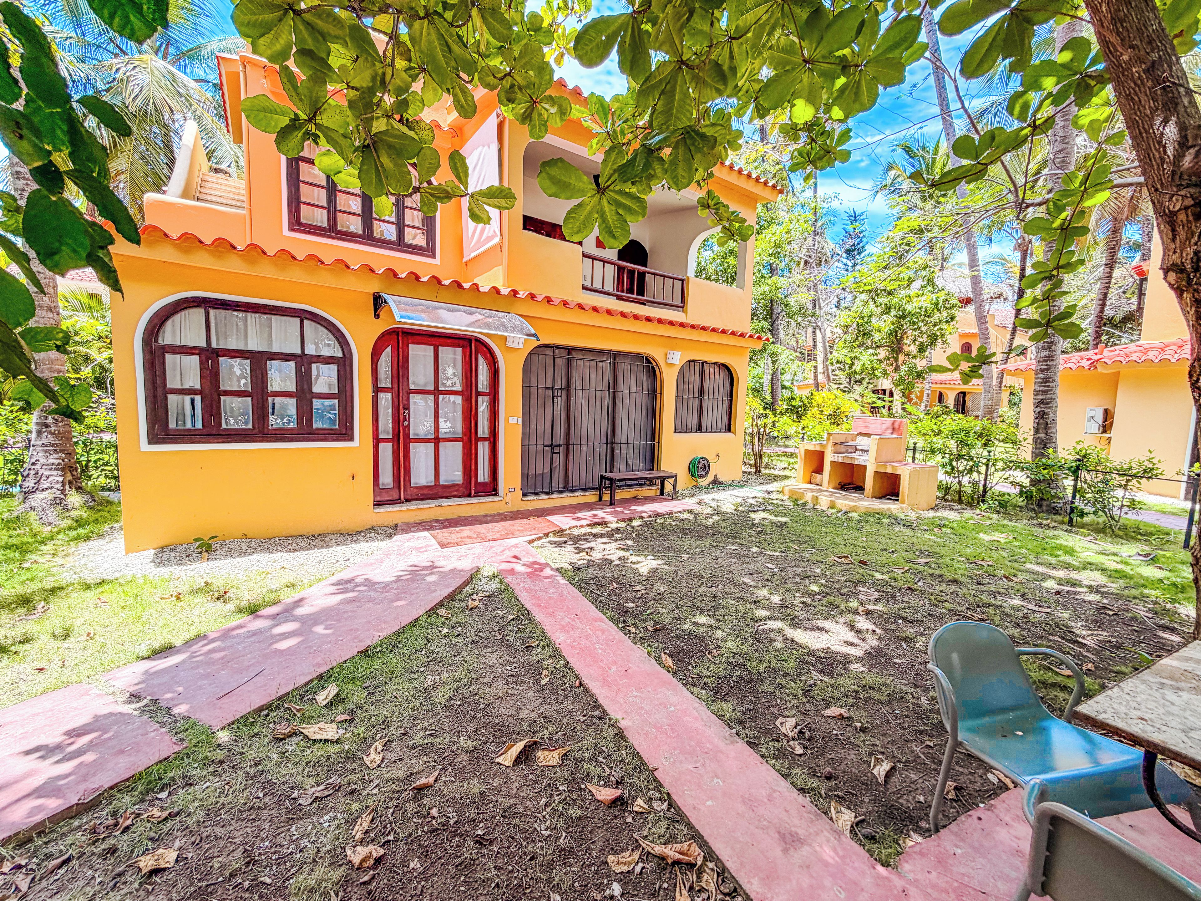 Un edificio de dos pisos con una puerta roja y varias ventanas, rodeado de follaje y un sendero pavimentado.