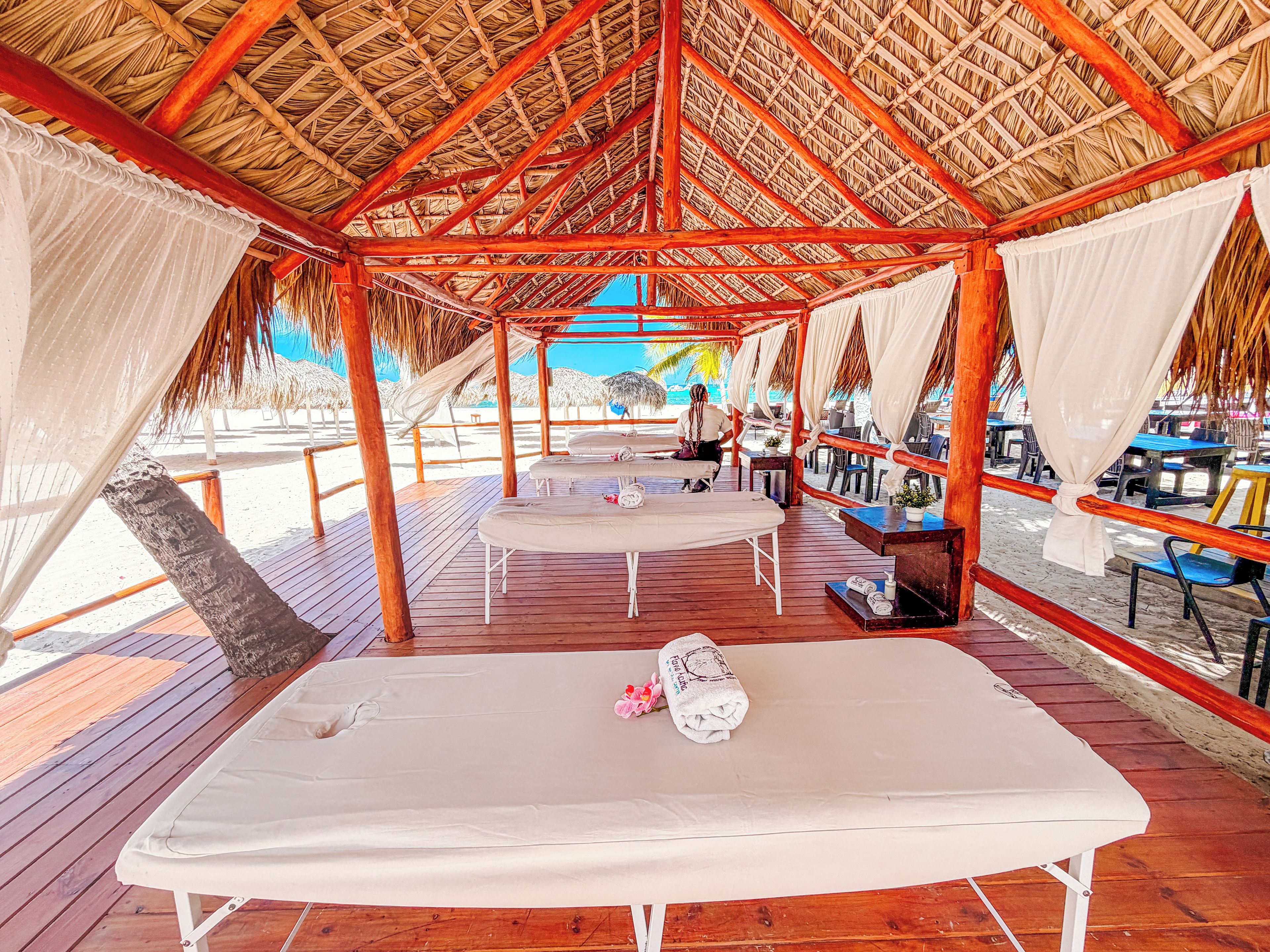 Una cabaña en la playa con techo de paja, terraza de madera y una mesa de masaje.