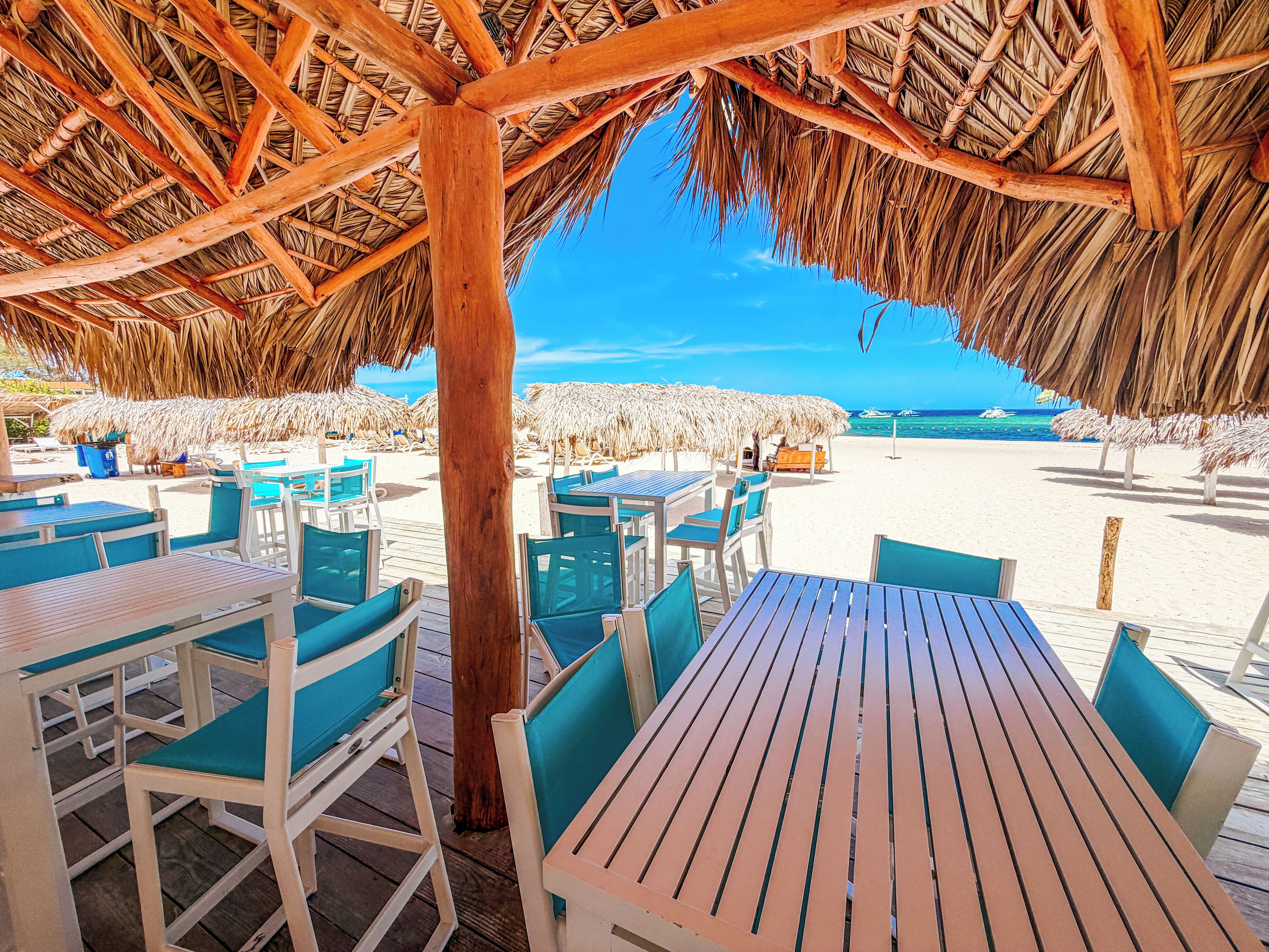 An outdoor seating area with wooden tables and chairs, a thatched roof, and a view of the beach and ocean.