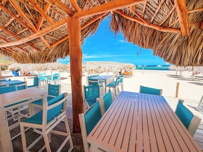 An outdoor seating area with wooden tables and chairs, a thatched roof, and a view of the beach and ocean.