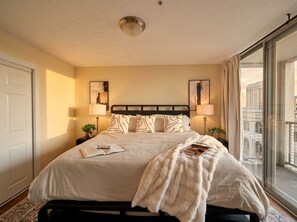 A hotel room with a large bed, two bedside lamps, a window with a view, and a door. - Prime Cleveland Location  Modern 2BR + Balcony (Cleveland)