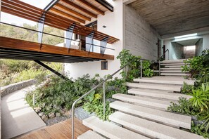 Un escalier moderne avec des marches en béton et des rampes en métal, menant à une terrasse en bois et à un balcon offrant une vue sur la verdure environnante.