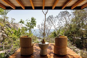 Une terrasse en bois avec deux chaises en osier et une table centrale, offrant une vue sur un paysage côtier avec des arbres et un ciel dégagé.