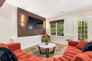 A living room with a large flat-screen TV mounted on a patterned wall, a round coffee table, and orange sofas. - 7 Kings / Hot Tub + Sauna + Game Room / Sleeps 20 (Massanutten)