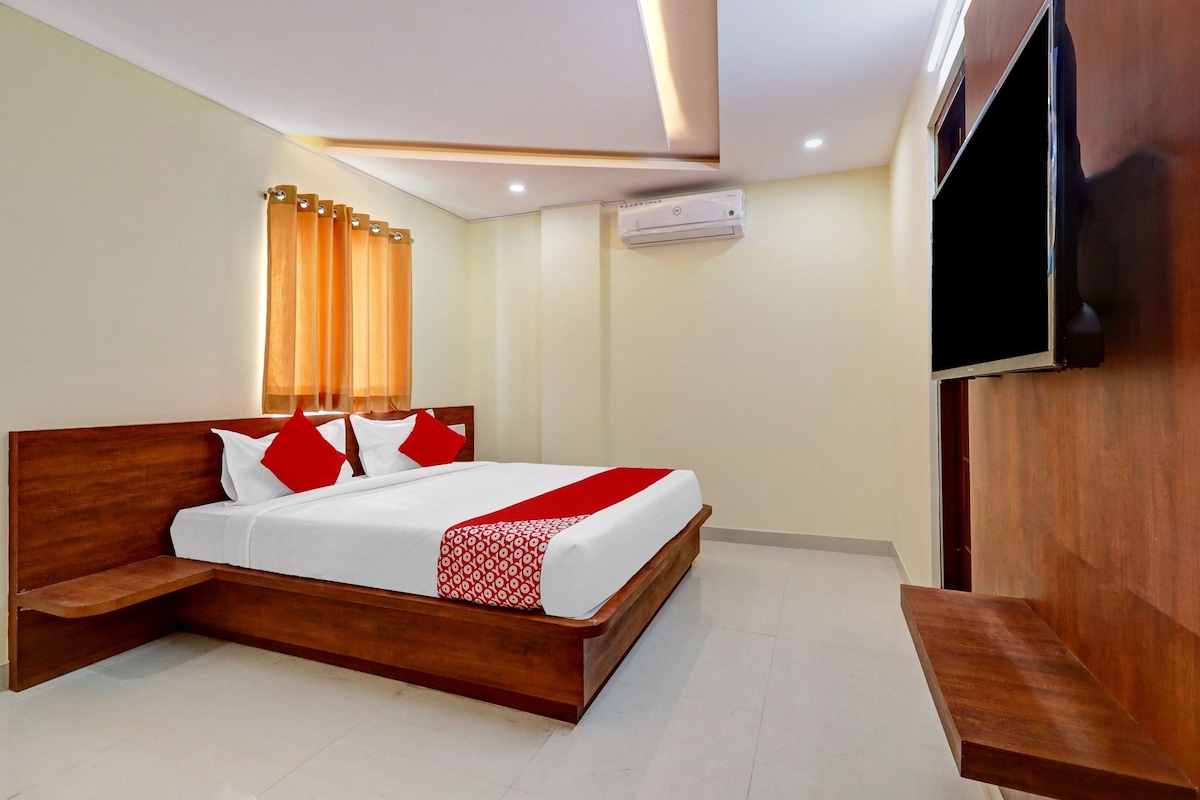 Quarto de hotel com cama de madeira, roupa de cama branca, almofadas vermelhas, manta estampada em vermelho e branco, TV de tela plana presa na parede e aparelho de ar condicionado.