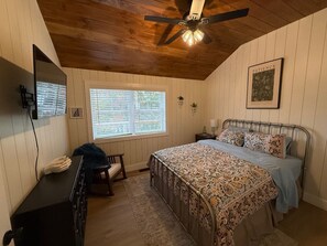 Ein Schlafzimmer mit einem Bett, Deckenventilator, Fenster mit Jalousien, Nachttisch und einem an der Wand befestigten Fernseher.