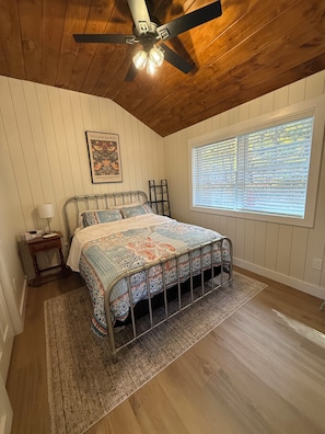 A bedroom with a bed, a ceiling fan, a window with blinds, a framed picture on the wall, and a small bedside table. -   The serenity of a treehouse with  access to a beautiful lake (Stoddard)