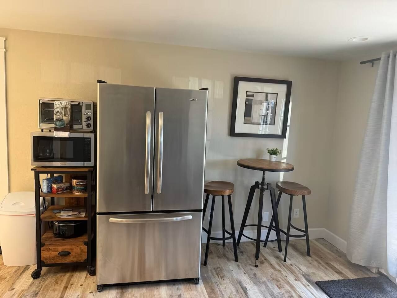 A compact kitchen with a stainless steel refrigerator, a microwave, and a small coffee machine on a shelf. There are two stools and a small table with a plant.