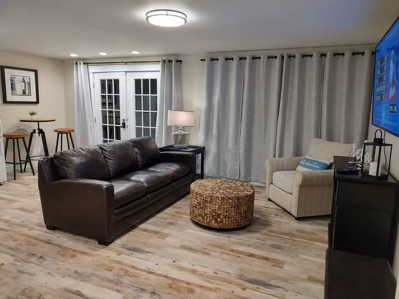 A living room with a leather sofa, a round ottoman, a side table with a lamp, a television, and a window with curtains.