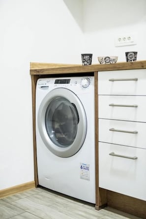 Un lave-linge compact, encastré, doté d’un panneau de commande, d’une étagère en bois au-dessus et d’une armoire blanche avec des tiroirs sur sa droite.