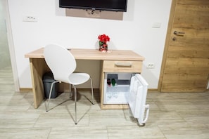 Une pièce de taille modeste, comprenant un bureau en bois, une chaise blanche, un petit réfrigérateur et un téléviseur fixé au mur.