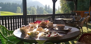 Une table ronde, dressée pour le petit-déjeuner, entourée de chaises en osier, sur un balcon offrant une vue sur des champs verdoyants et des montagnes.