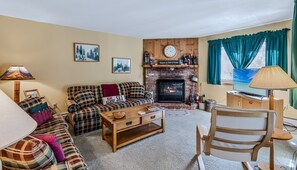 A cozy living room with a fireplace, plaid sofa, and a television. - Kettlebrook Ski-In/Ski-Out Condo with Fireplace (Ludlow)