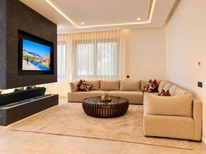 Una sala de estar moderna con un televisor de pantalla plana montado en una pared oscura, un sofá seccional color beige, una mesa de centro redonda y un amplio ventanal con cortinas traslúcidas.
