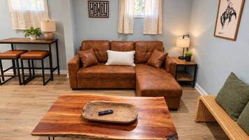 Un salon comprenant un canapé d’angle en cuir marron, une table basse en bois, un canapé en osier, une petite table avec une lampe et un tableau encadré accroché au mur.