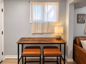 Un petit coin repas avec une table et deux chaises, une lampe sur la table et un canapé à l’arrière-plan.