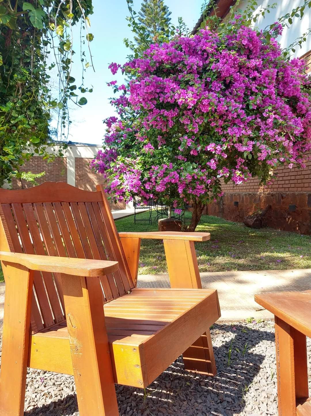A wooden outdoor seating area with a bench and a table, surrounded by a garden with a flowering bush.
