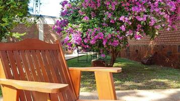 A wooden outdoor seating area with a bench and a table, surrounded by a garden with a flowering bush.