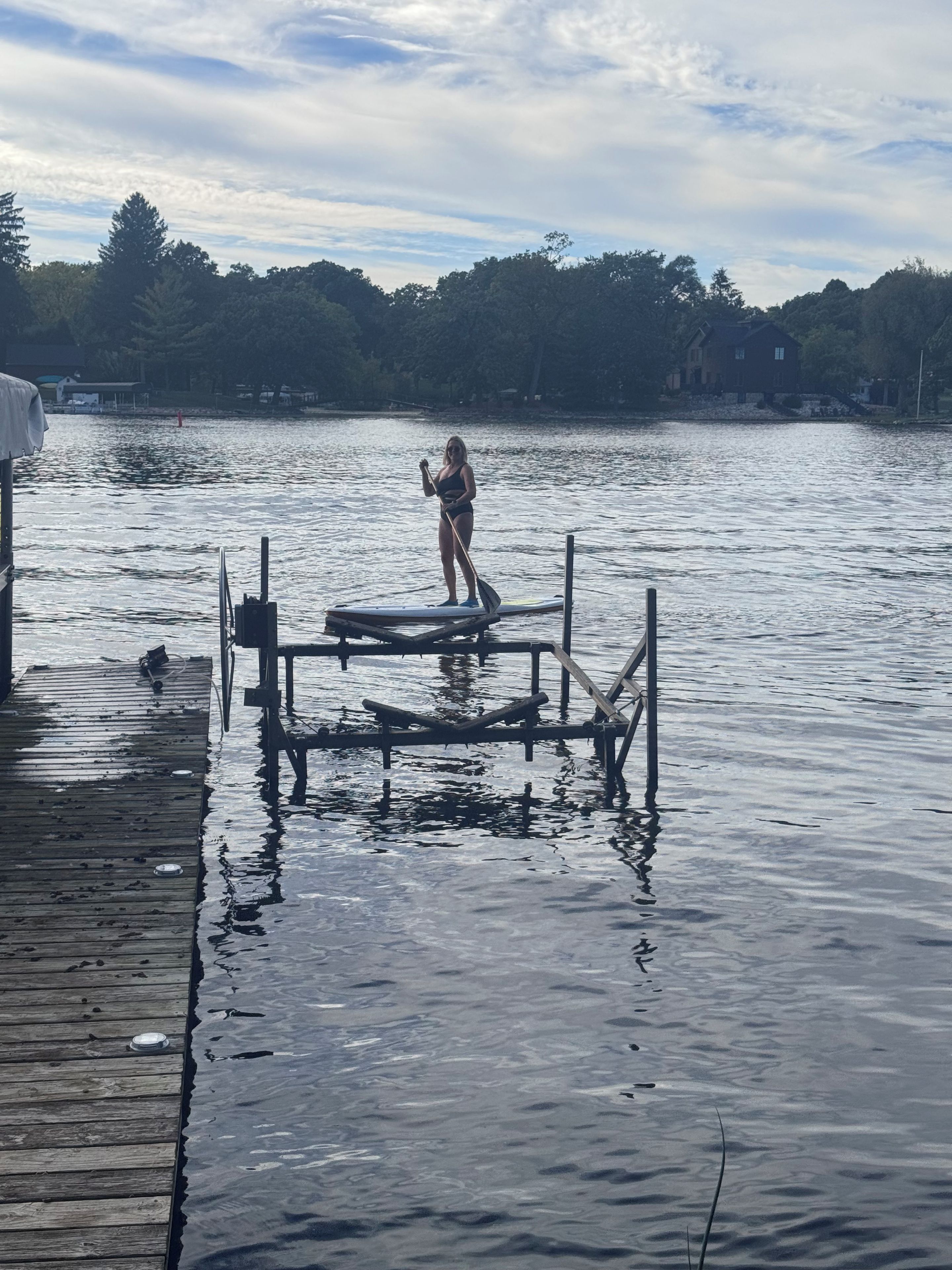 A person standing on a dock with a paddleboard, surrounded by calm water and a dock extending into the water.