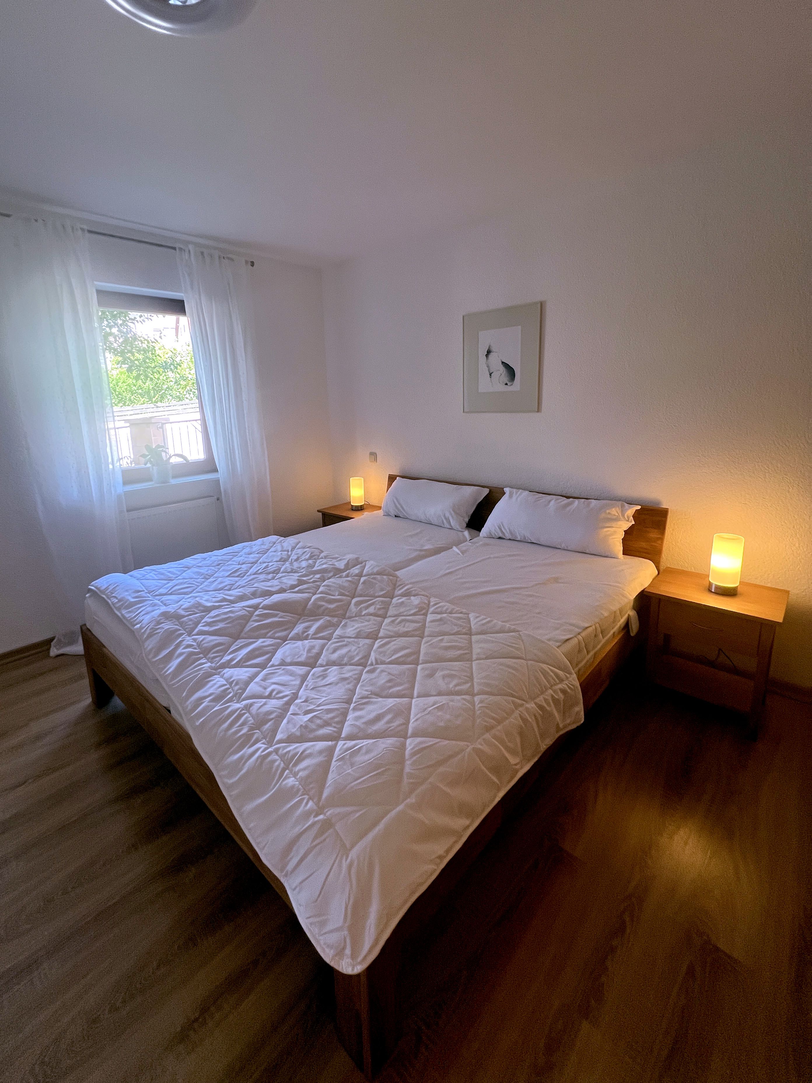 Ein ordentlich bezogenes Bett mit einer gesteppten weißen Decke, zwei Nachttische mit Lampen, ein Fenster mit Vorhängen und ein gerahmtes Bild an der Wand.