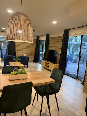 A modern living room with a wooden dining table, dark green upholstered chairs, a woven pendant light, a flat-screen TV on a wooden stand, and a brick wall. - Beautiful desert gem townhouse dual masters in North Scottsdale (Scottsdale)