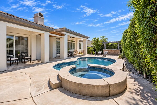 A swimming pool with a curved design, a patio area with outdoor furniture, and a well-maintained hedge. - Desert Dreamin' with Private Pool & Spa (Palm Desert)