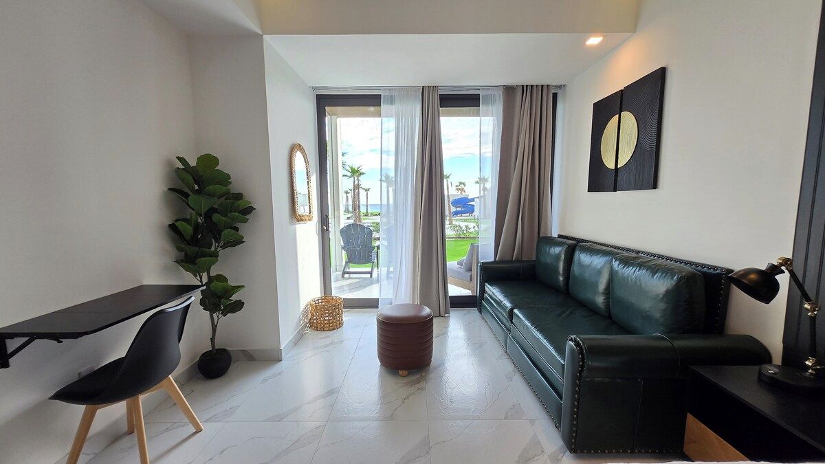 Un salon moderne avec un canapé en cuir noir, un bureau noir avec une chaise, une plante en pot et une vue sur l’extérieur grâce à une porte vitrée.