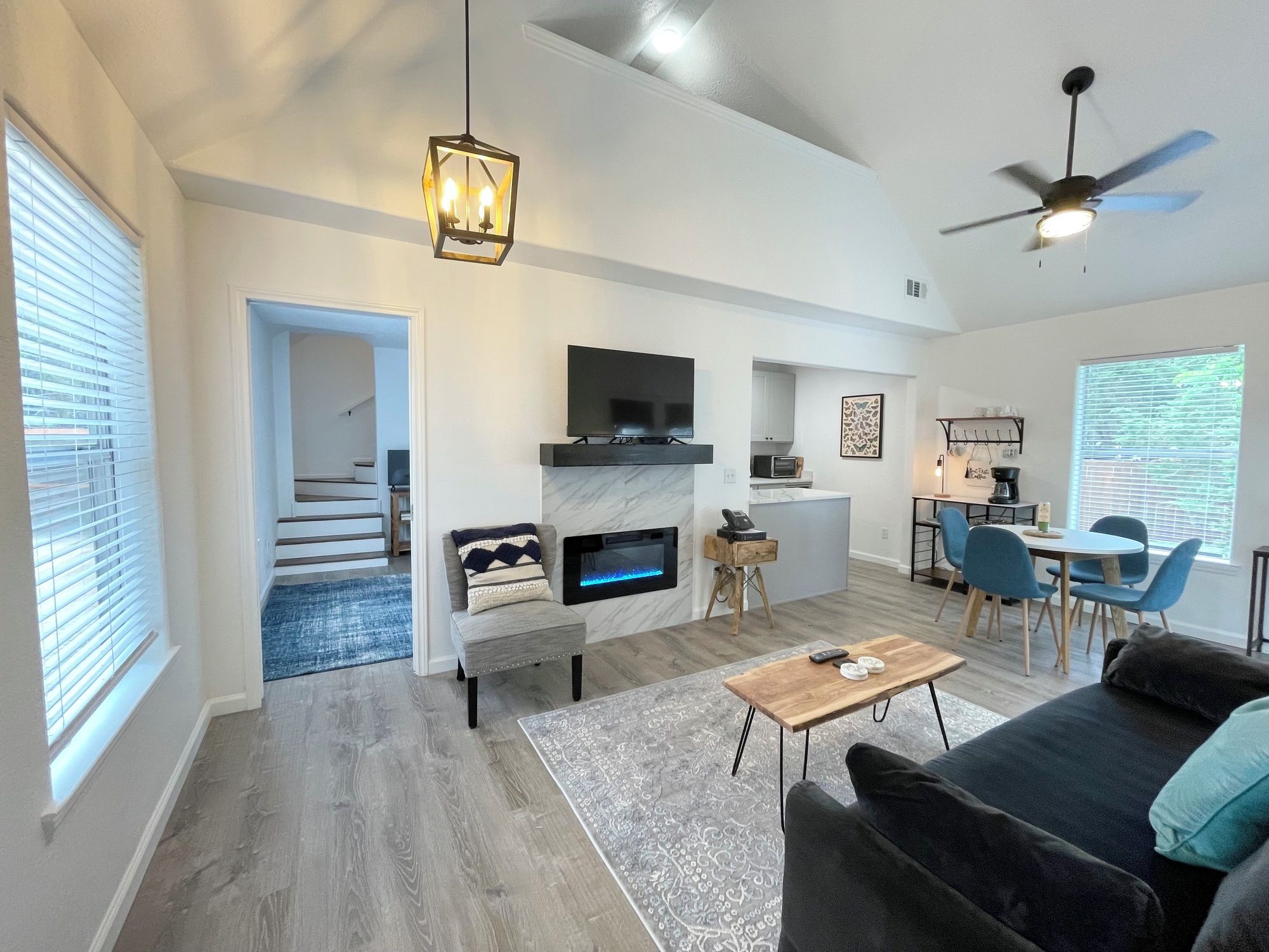 A modern living room with a fireplace, a flat-screen TV, a ceiling fan, and a dining area with blue chairs.