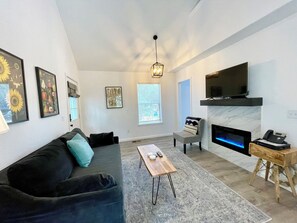 A modern living room with a sofa, a coffee table, a fireplace, and a TV mounted on the wall. - Cottage 7 Blue, Pet-Friendly One-Bedroom Cottage with Loft, Hot Tub (Wimberley)