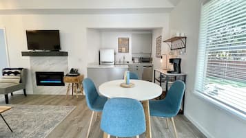 A modern living room with a fireplace, a dining area with blue chairs, a kitchen with white cabinets, and a large window with blinds.
