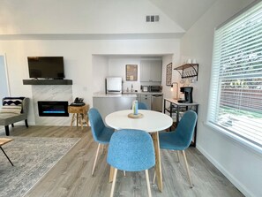 A modern living room with a fireplace, a dining area with blue chairs, a kitchen with white cabinets, and a large window with blinds. - Cottage 7 Blue, Pet-Friendly One-Bedroom Cottage with Loft, Hot Tub (Wimberley)