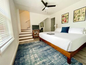 Un dormitorio con una cama, ventilador de techo, televisor, escritorio y una escalera que conduce a un nivel superior.