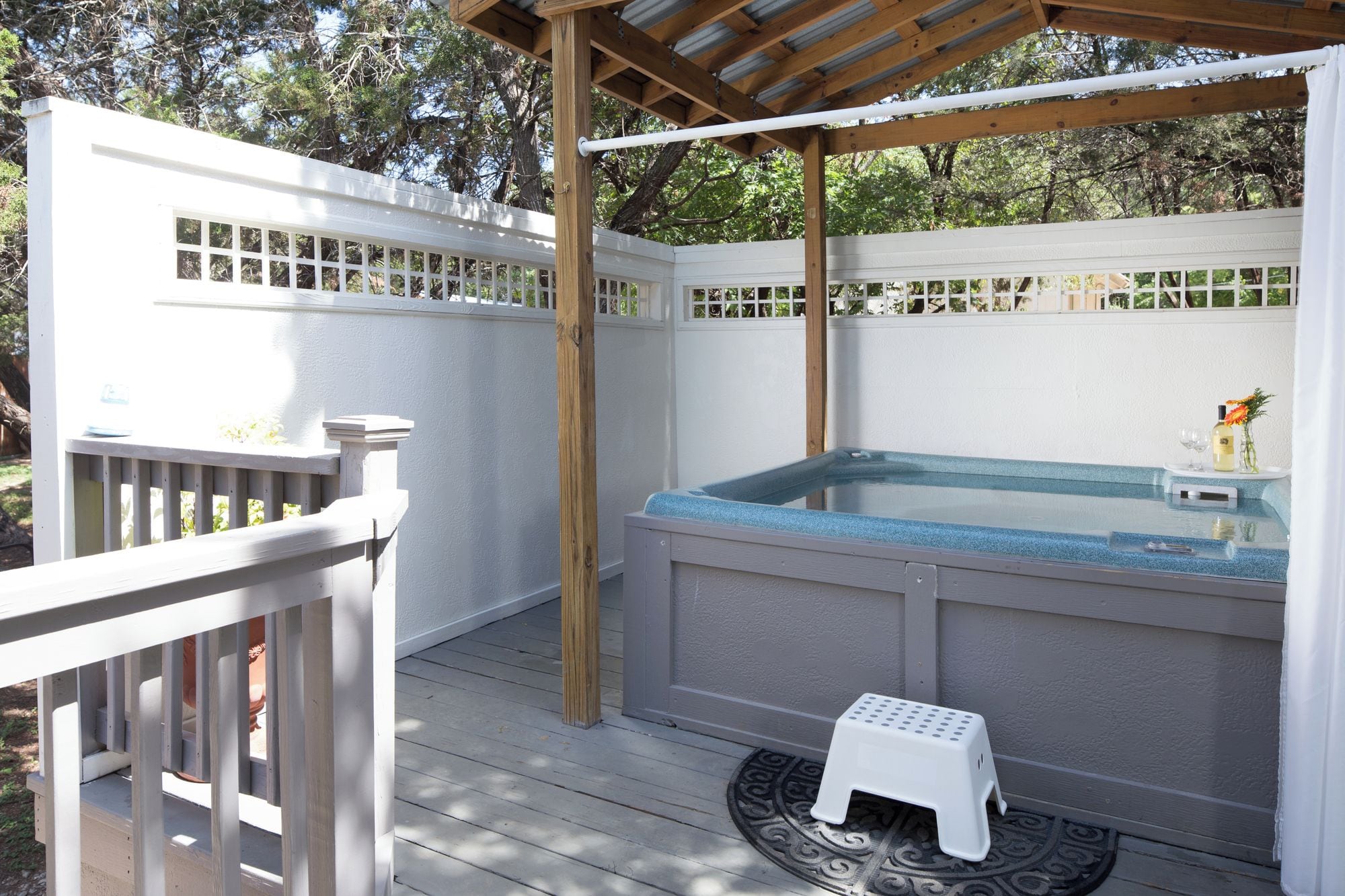 A hot tub under a wooden pergola with a white fence and a small white stool.
